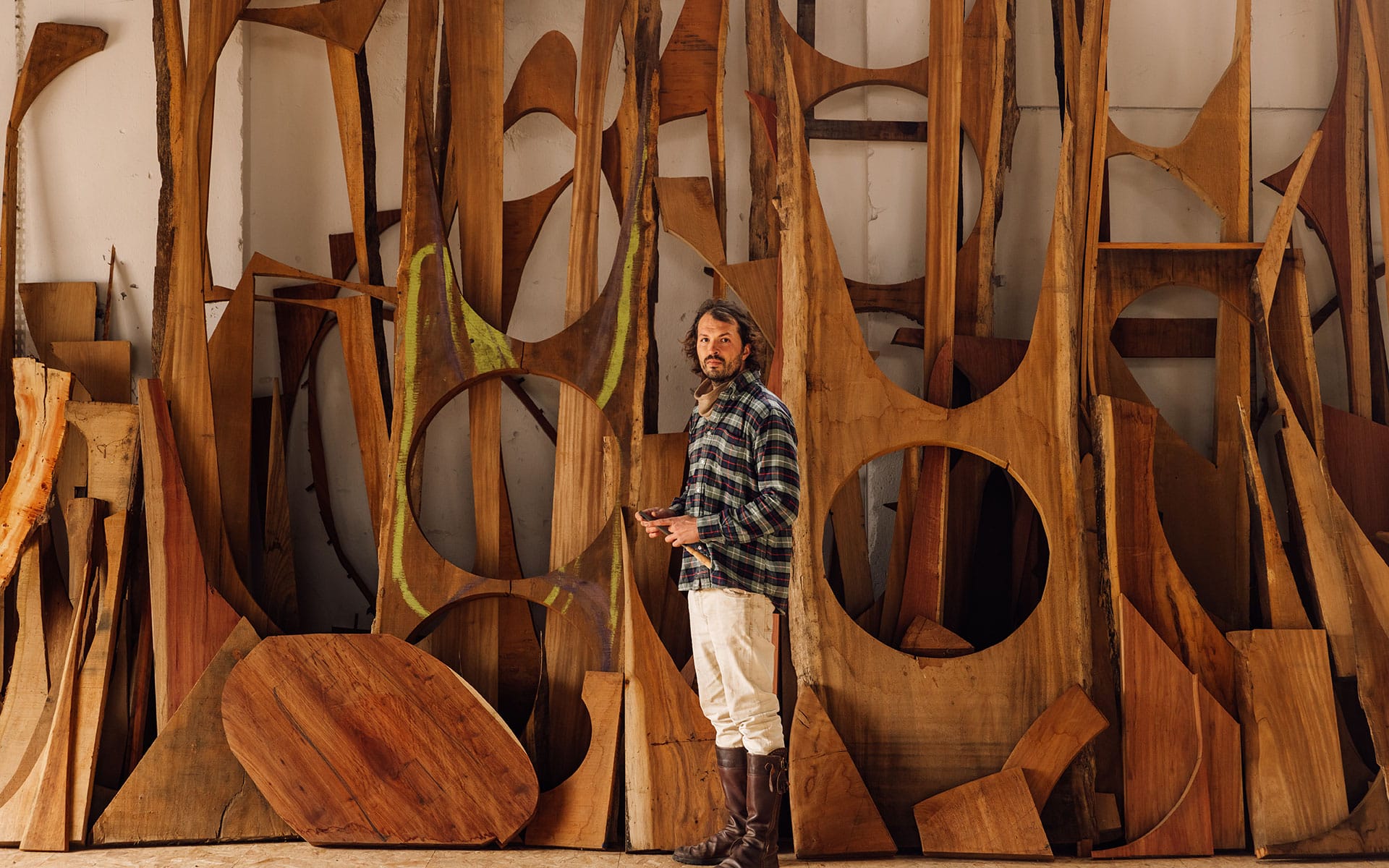 Man in a workshop surrounded by large wooden sculptures and abstract shapes, wearing a plaid shirt and boots.