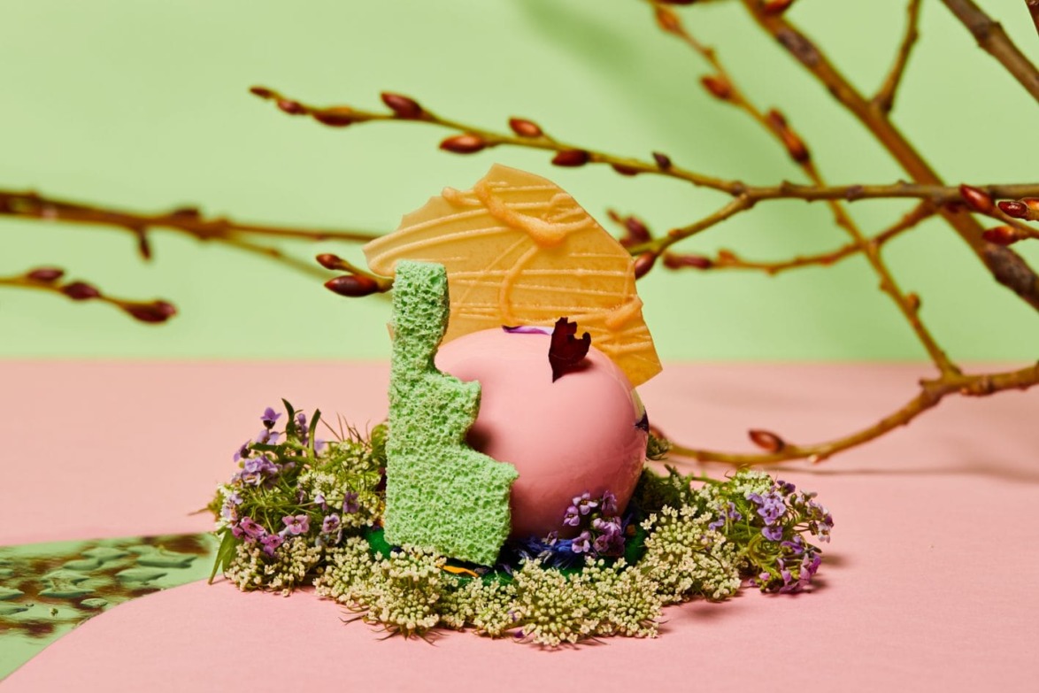 Pink dessert with green and yellow decorations, surrounded by small flowers and branches on a pastel background.