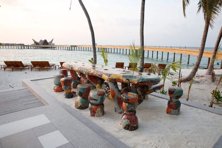 Colorful mosaic table with matching stools on sandy beach, overlooking pier and ocean, surrounded by palm trees.