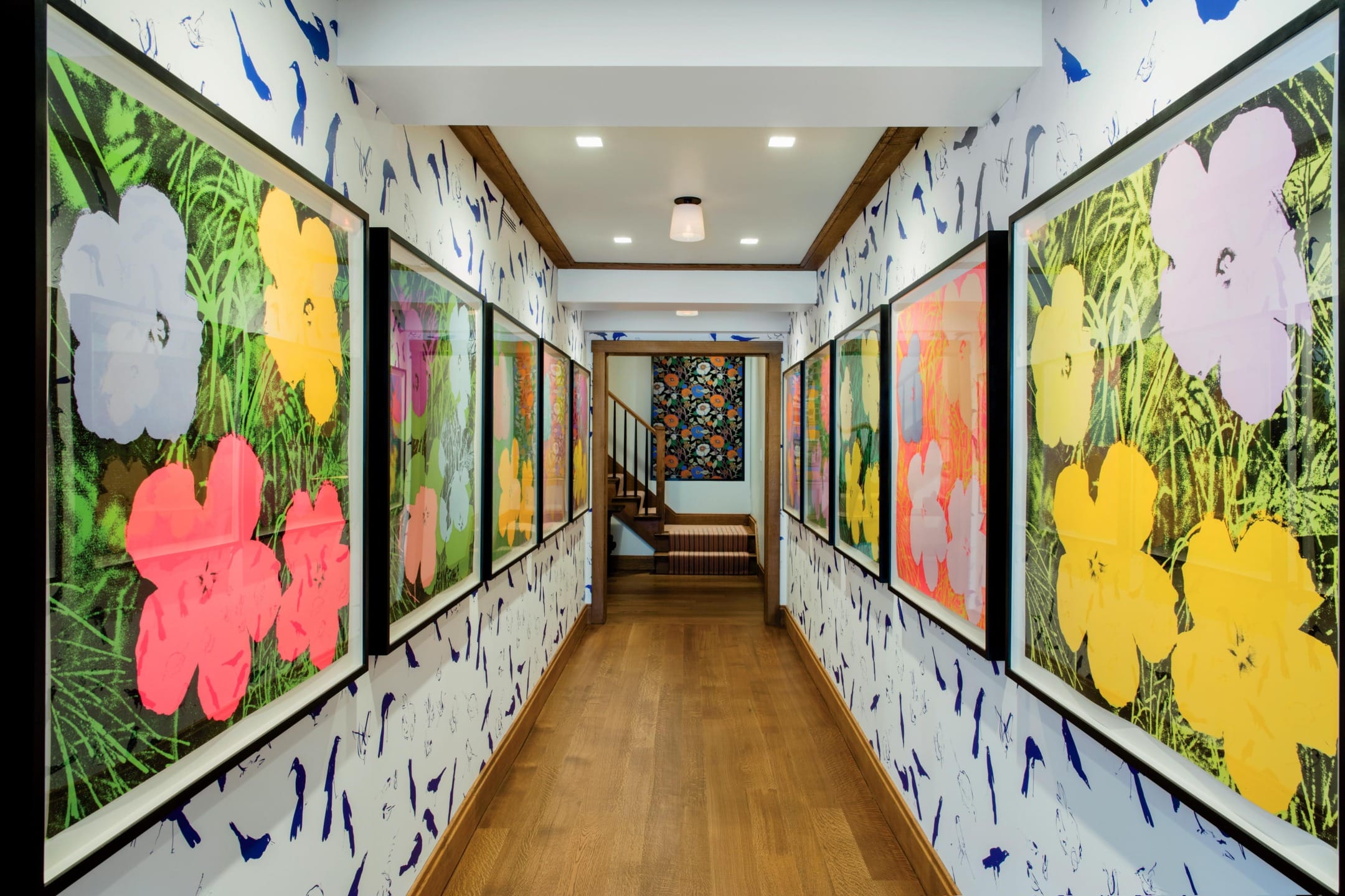 Hallway with colorful framed floral artwork on both sides, wooden floor, and stairs leading to another room.