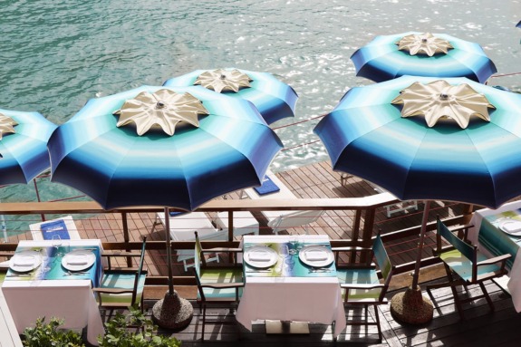 Riverside dining area with blue umbrellas over tables set for two by the water.