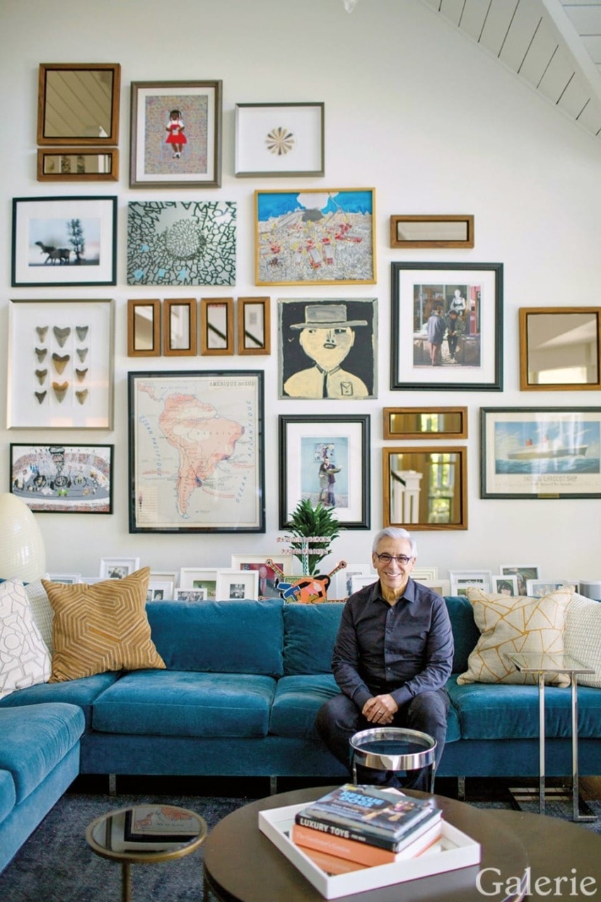 Man seated on a blue sofa in a living room with a gallery wall filled with various framed artworks and photographs.