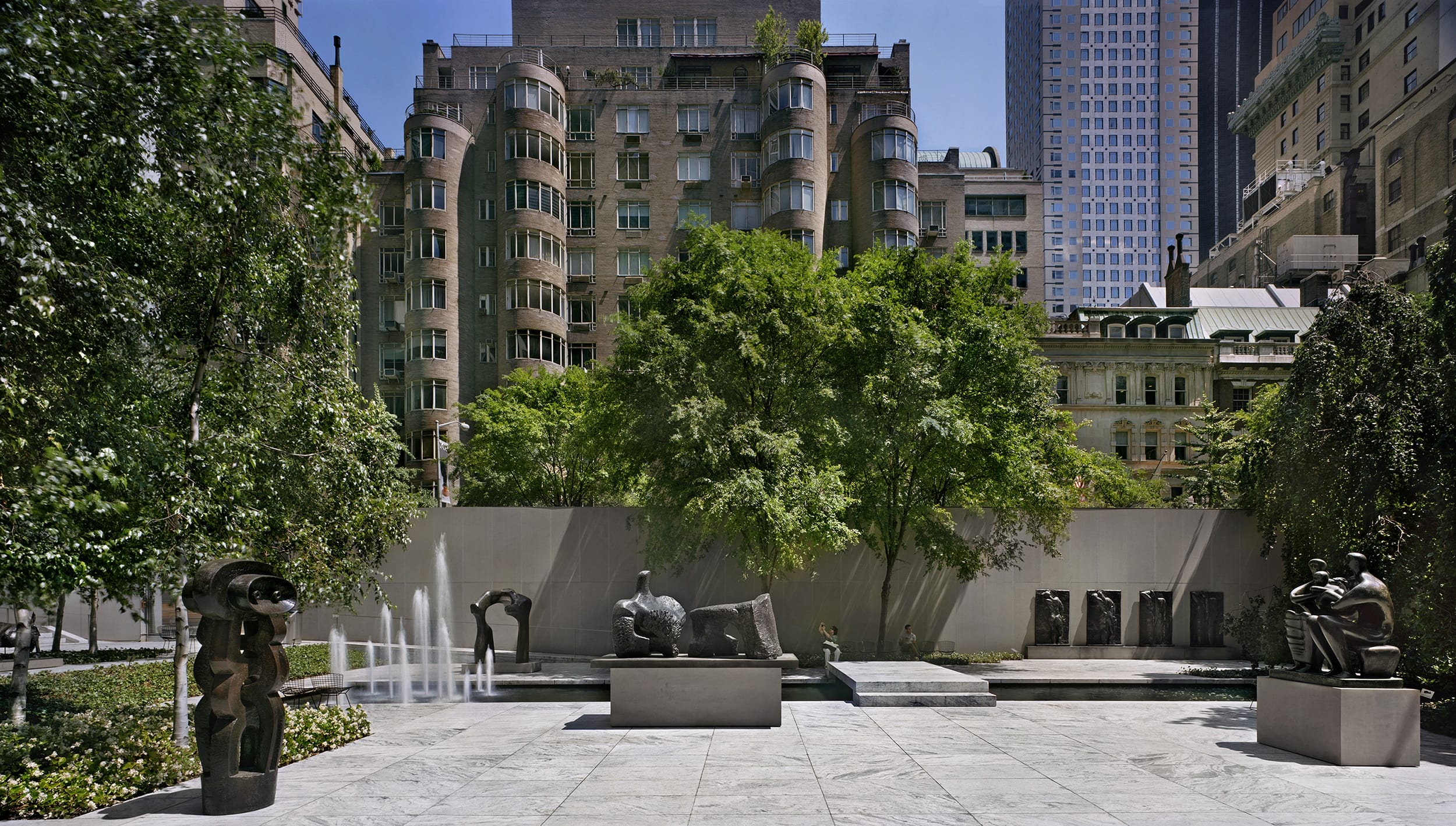 Outdoor sculpture garden with modern art, trees, and urban buildings in the background on a sunny day.