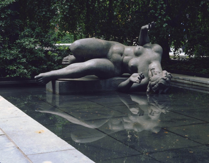 Statue of a reclining figure in a fountain surrounded by greenery, reflecting in the water below.