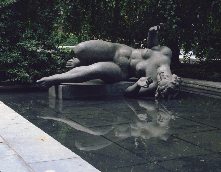 Statue of a reclining figure in a fountain surrounded by greenery, reflecting in the water below.