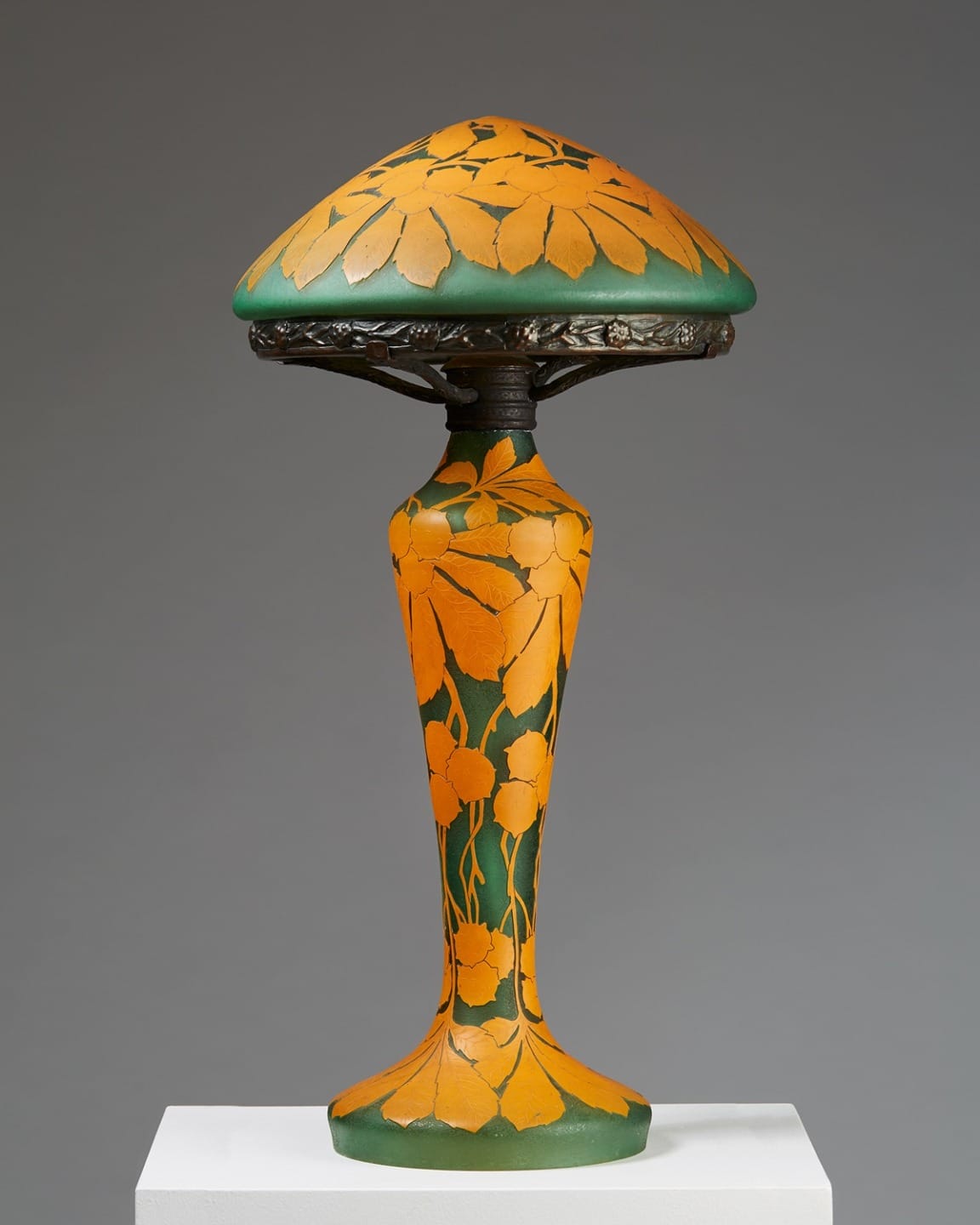 Art nouveau style lamp with orange floral pattern on green base and shade, displayed on a white pedestal against a gray background