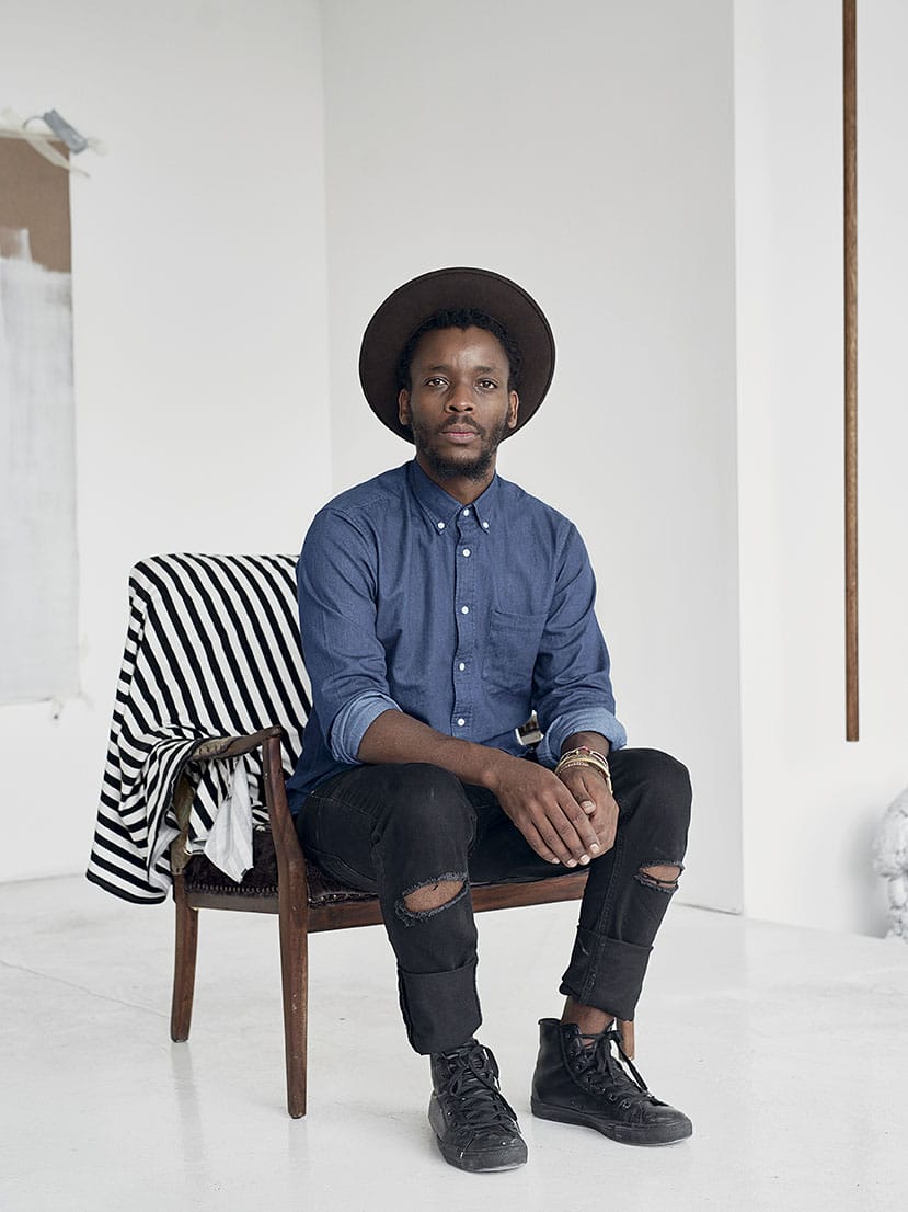 Man sitting on a chair wearing a hat, blue shirt, and black pants, with a striped fabric draped over the chair.