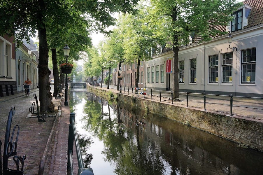 Canal view in a historic European town with tree-lined streets, old buildings, and a small bridge in the distance.
