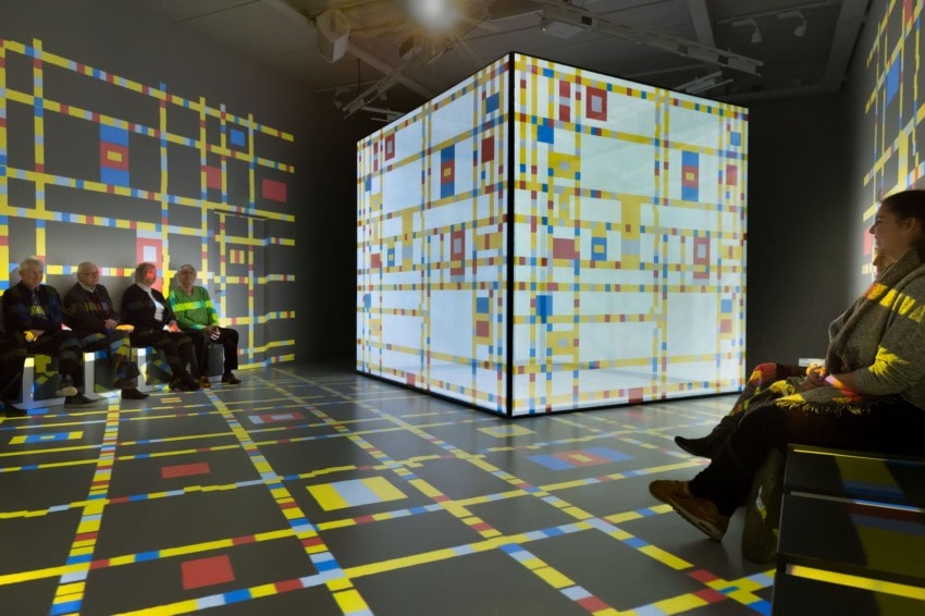Art installation with people observing a large cube displaying colorful geometric patterns projected on walls and floor.