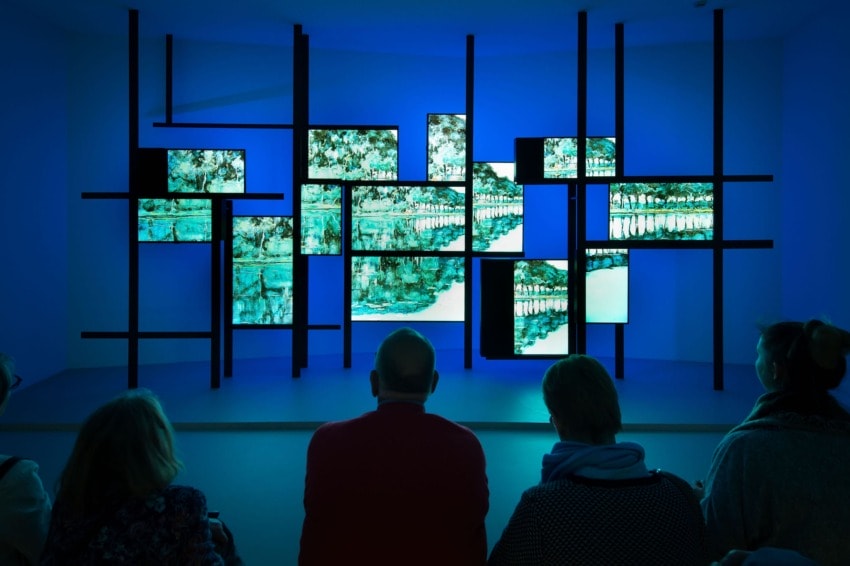 People sitting and watching a digital art display on a blue-toned wall with multiple screens.