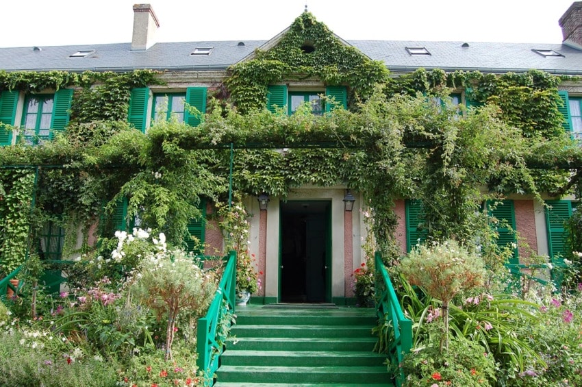 House covered in green ivy with matching shutters, surrounded by lush garden and stairs leading to entrance.