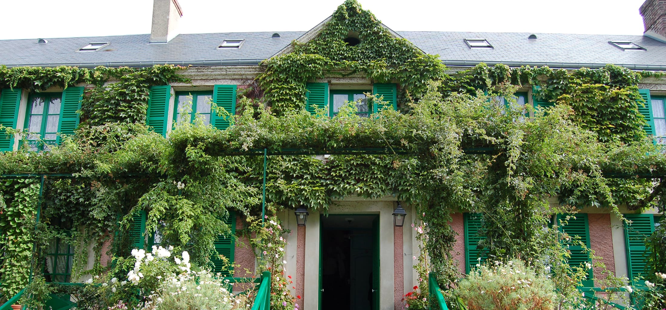 House with green shutters and ivy-covered walls, surrounded by lush garden flowers and greenery.