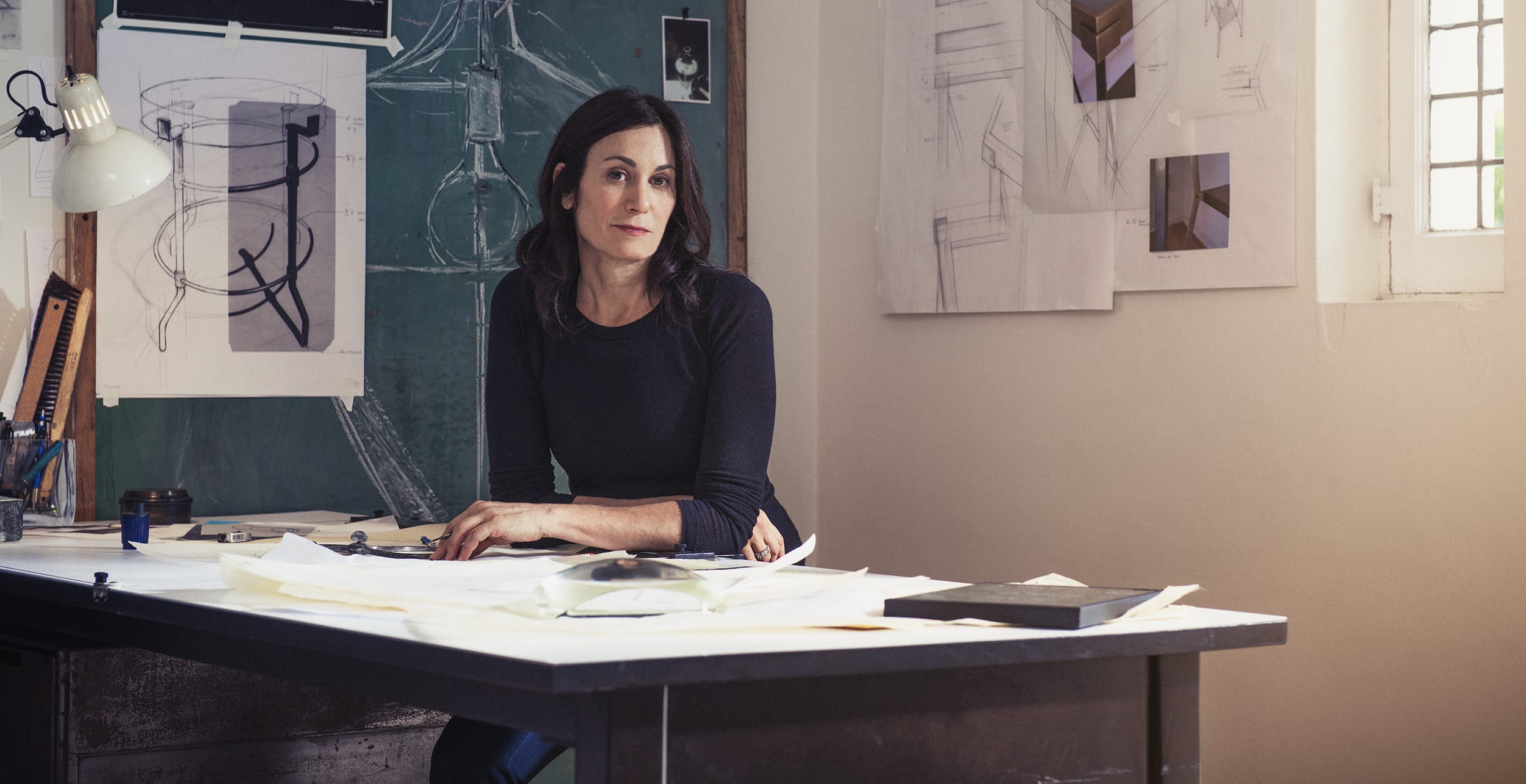 Person sitting at a design desk with sketches and architectural drawings on the wall in a well-lit room.
