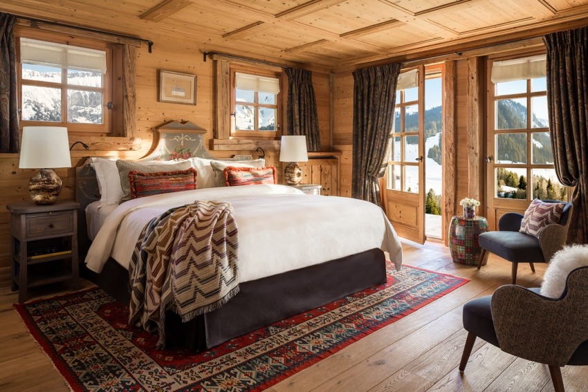 Cozy wooden alpine bedroom with large bed, patterned rug, and a scenic mountain view through glass doors.