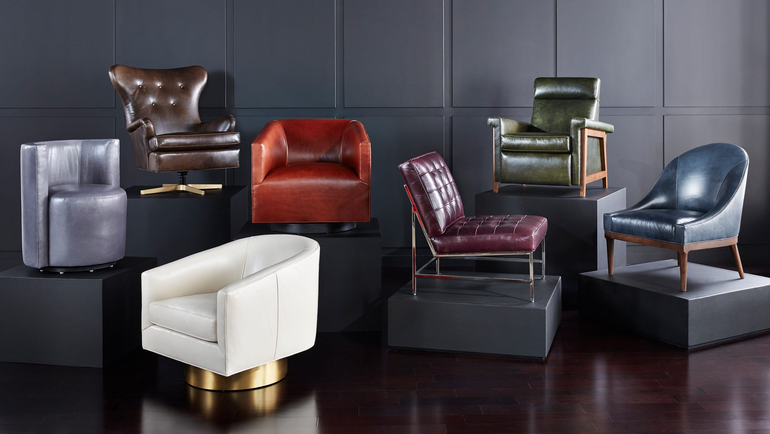 A variety of stylish leather chairs and armchairs displayed on dark platforms against a paneled wall background.