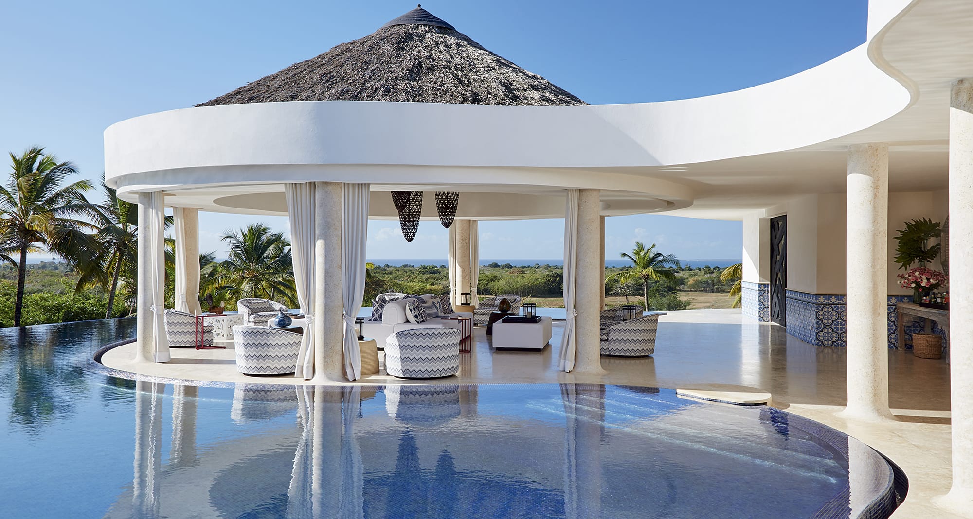 Luxury resort patio overlooking infinity pool with ocean view and palm trees in the background.