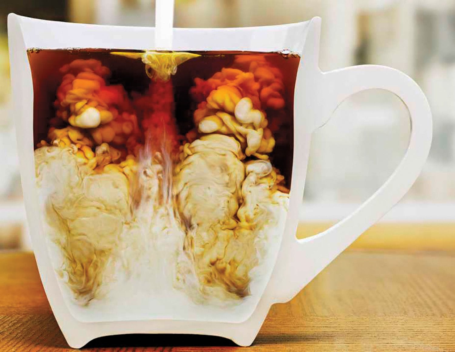 Cream being poured into a mug of black coffee, creating swirls of light and dark shades on a wooden table background.