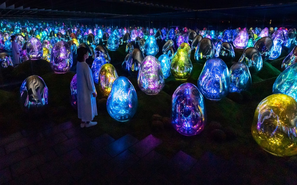Person observing vibrant, colorful illuminated sculptures in a dark room with reflections, creating a futuristic atmosphere.