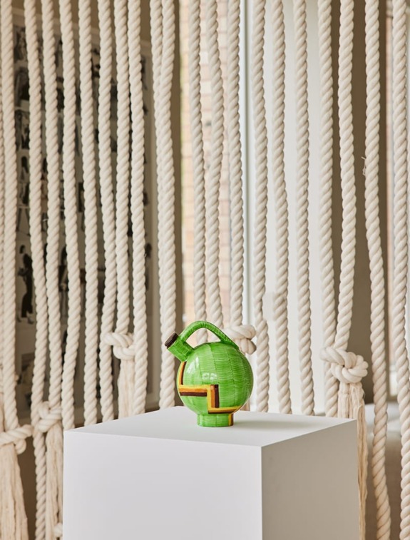 Green vase on white pedestal, background of large twisted rope art, minimalist interior design setting with soft lighting.