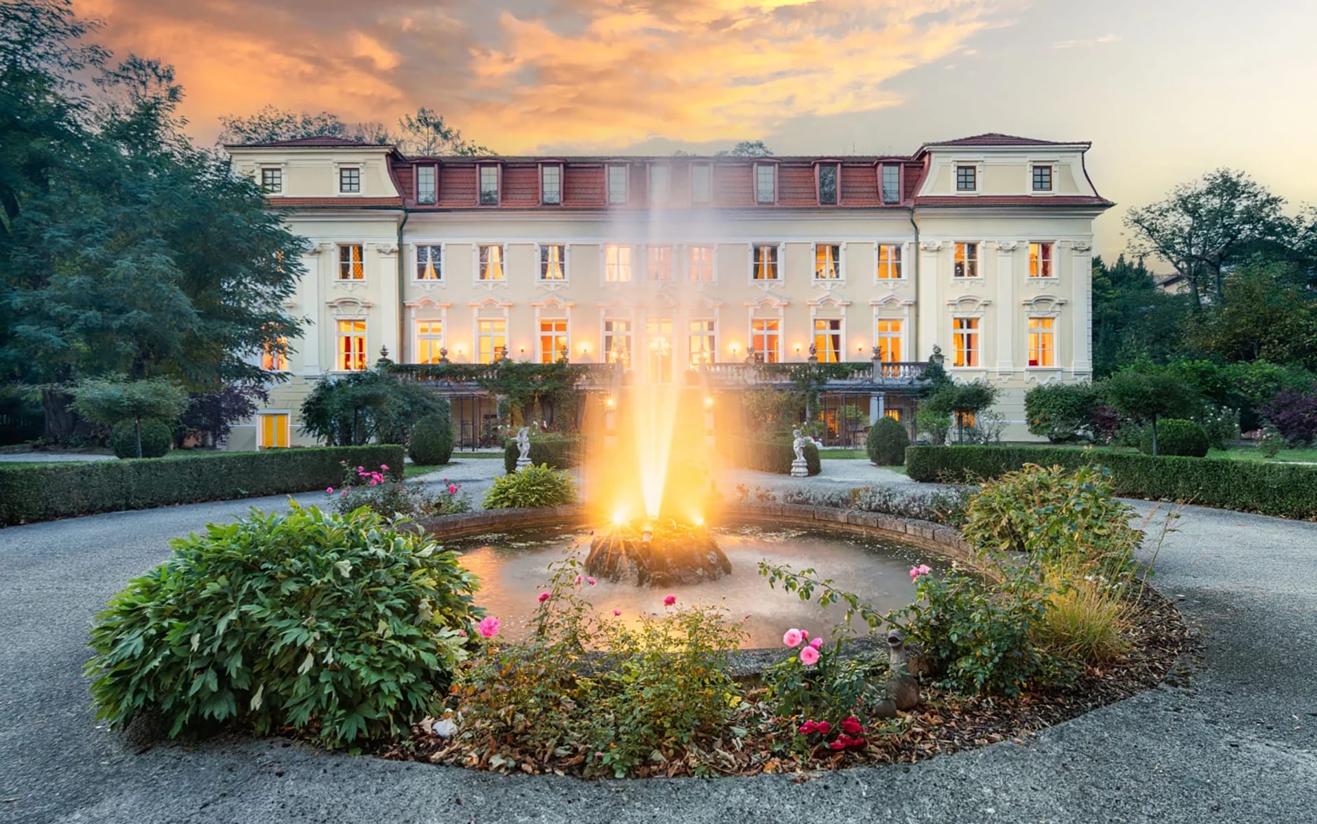 Grand historic mansion with illuminated fountain in front, surrounded by lush greenery and a vibrant sunset sky.
