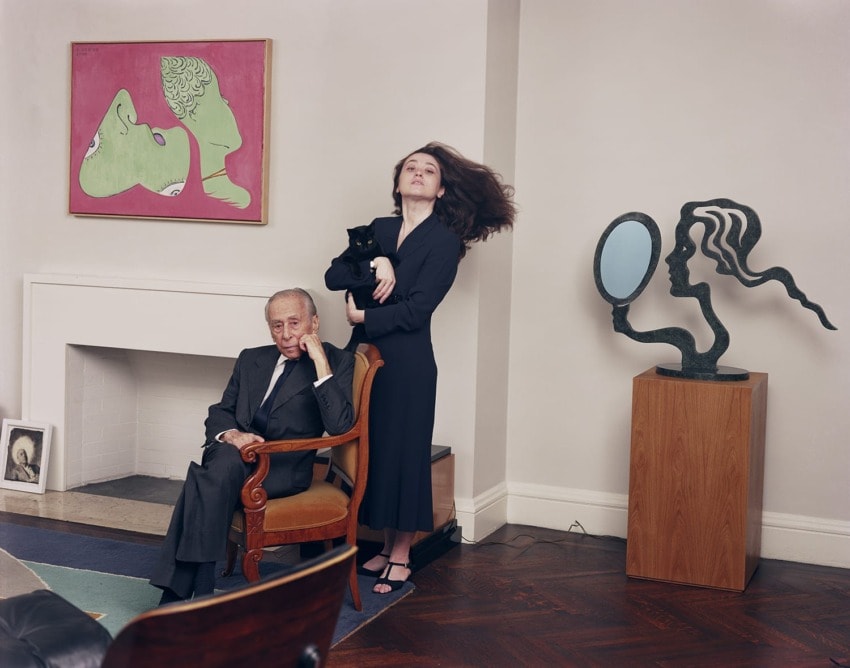 Man seated and woman holding a cat in an art-filled room with paintings and sculptures on display.