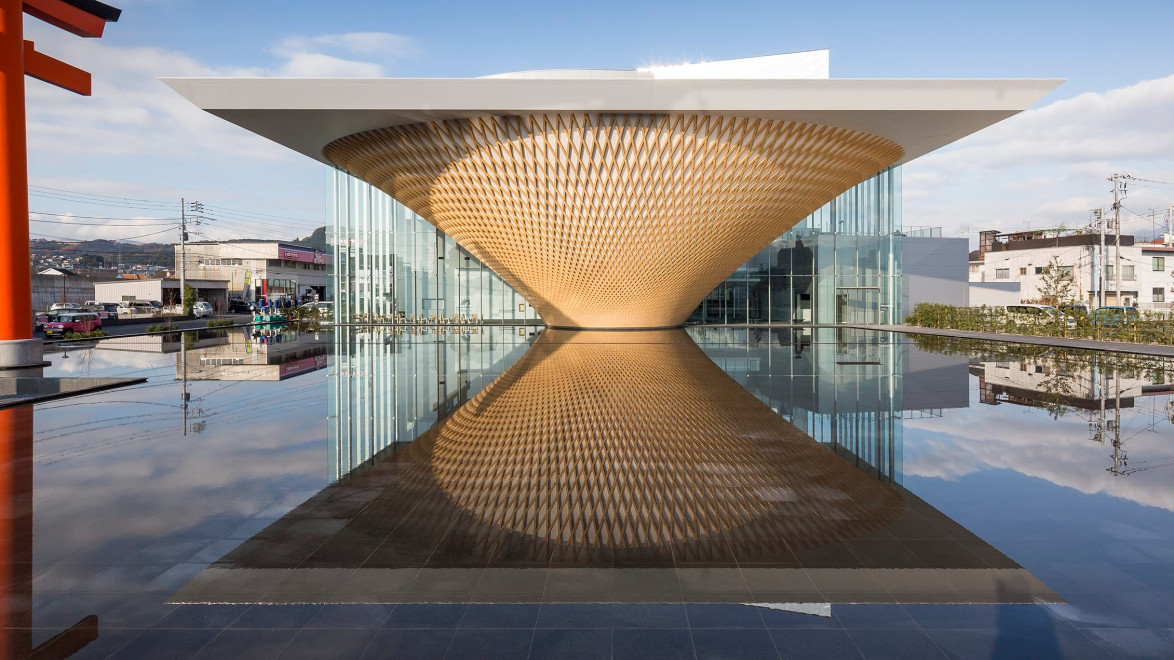 Modern building with a unique, curved facade and glass walls reflected in a still pool on a sunny day.