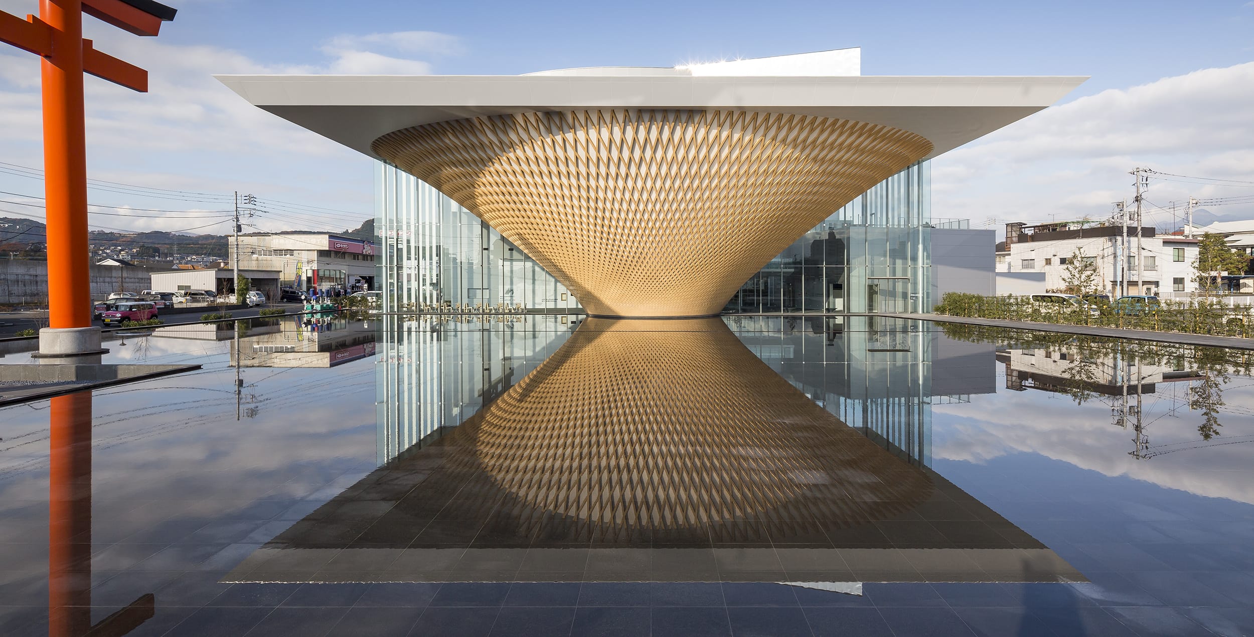 Modern building with a unique, curved facade and glass walls reflected in a still pool on a sunny day.