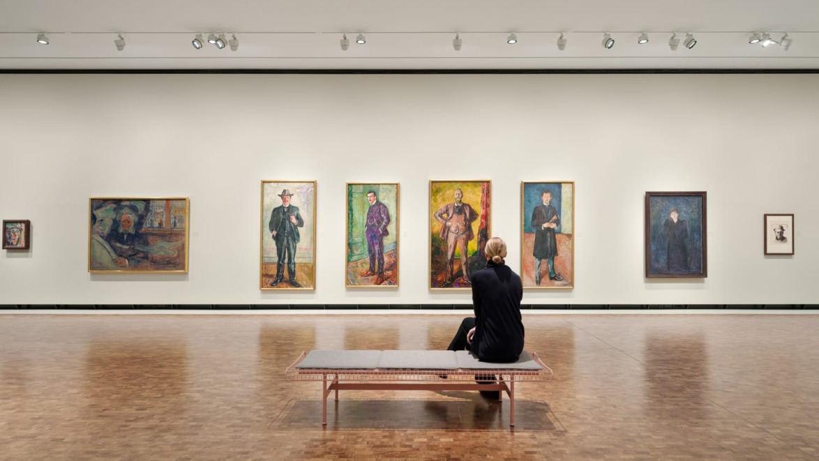 Visitor sitting on a bench in an art gallery, observing a row of portrait paintings on a well-lit wall.
