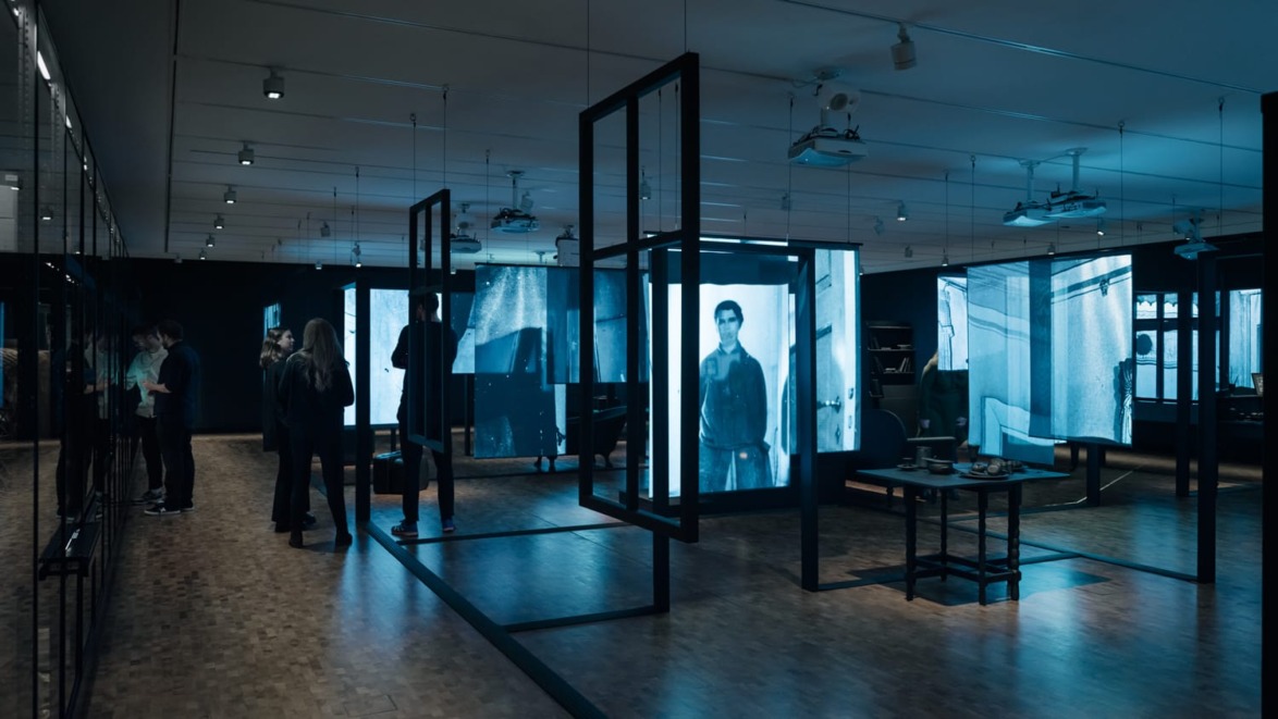 People observing bluish digital screens in a modern art gallery with ceiling projectors and wooden floor.