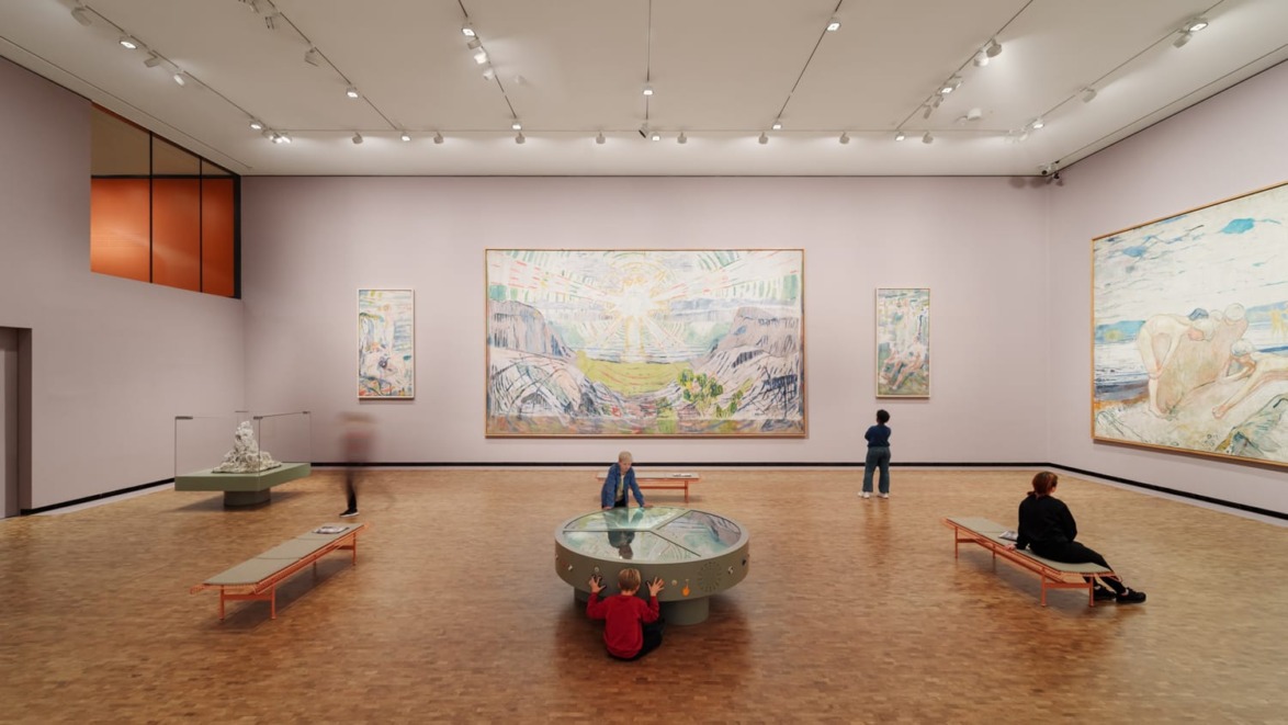 Visitors admire paintings in a spacious art gallery with large wall displays and seating benches.