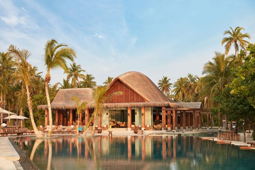 Tropical beach resort with a large pool, surrounded by palm trees and a thatched roof building under a clear blue sky.