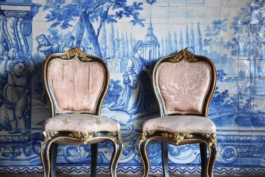Two ornate chairs with pink upholstery against a blue and white ceramic tile mural depicting historical figures and scenery.