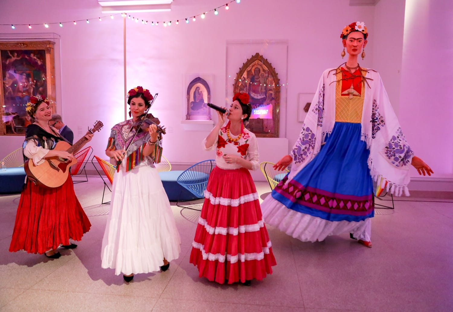 Performers in colorful traditional attire, one playing guitar, another singing, with a tall figure in a vibrant dress beside them.