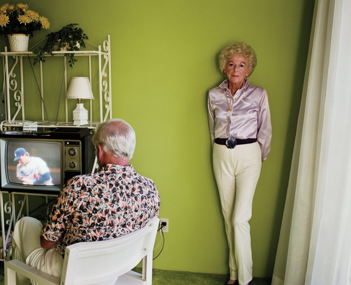 Elderly woman standing in a room with green walls while a man watches TV.