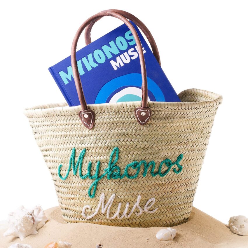 Woven beach bag with handles, embroidered with "Mykonos Muse," on sand with seashells, holding a blue book.