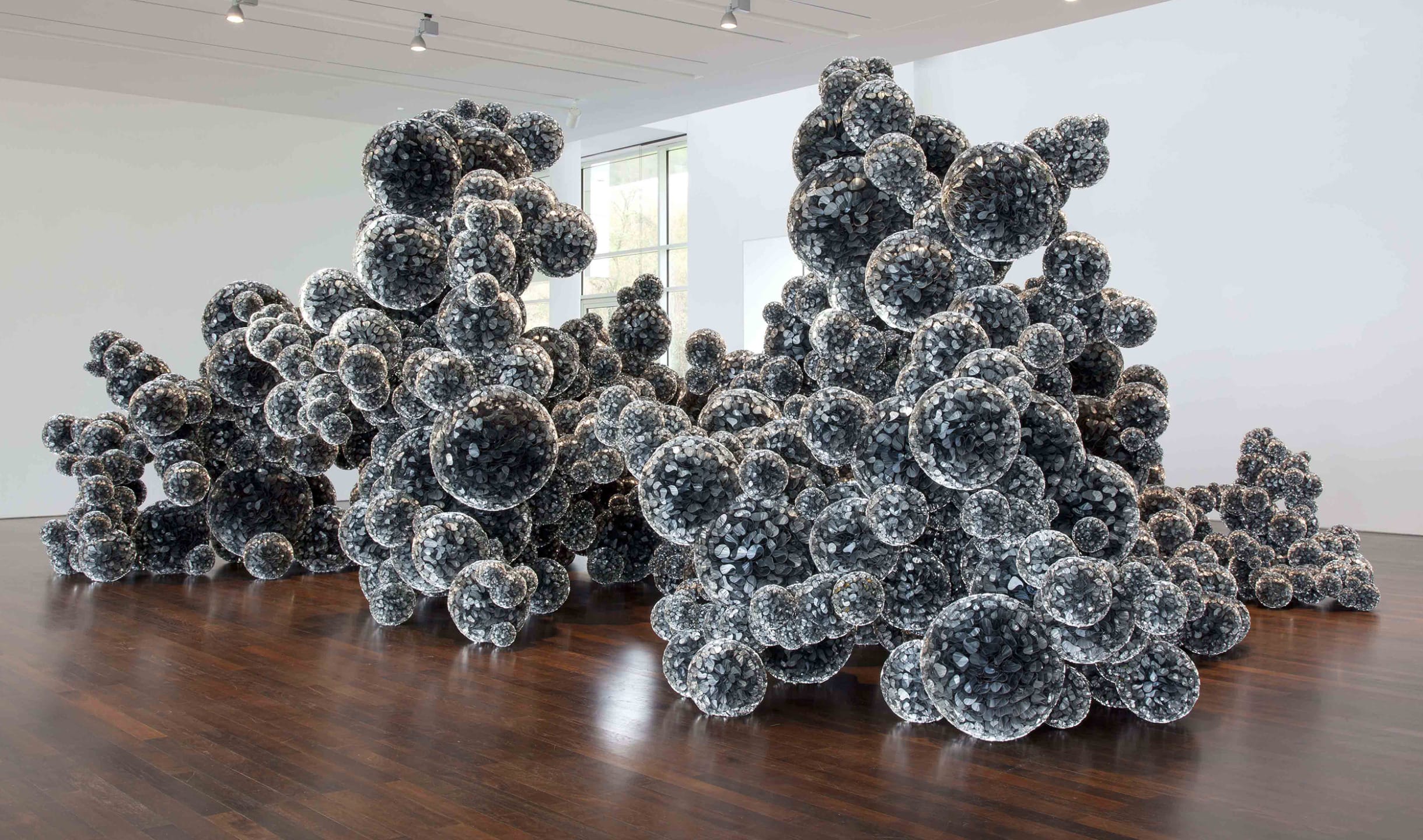 Sculpture composed of clusters of dark, reflective spheres displayed in a contemporary art gallery setting.