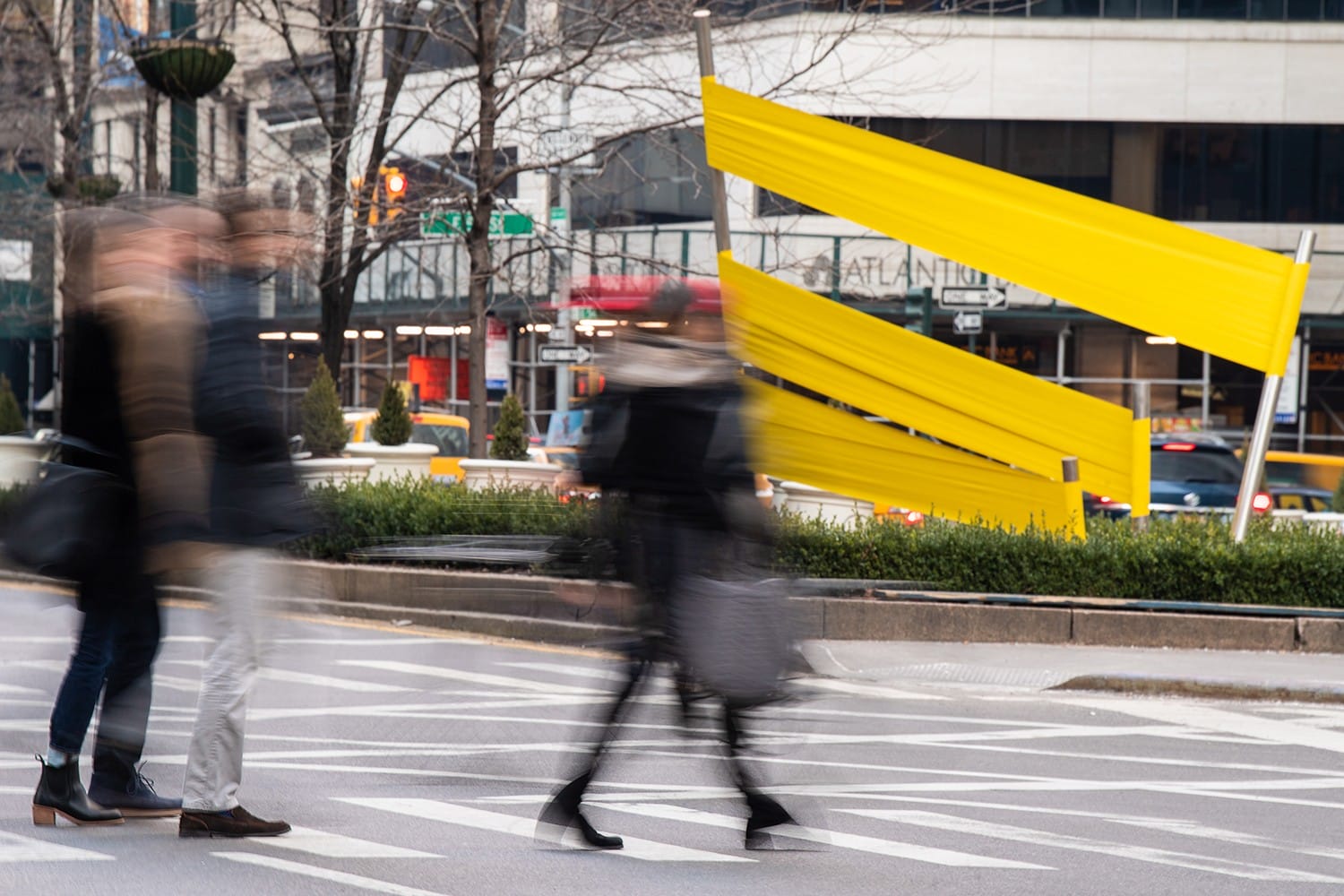 People walking on a city street with a yellow art installation in the background, capturing motion and urban life.