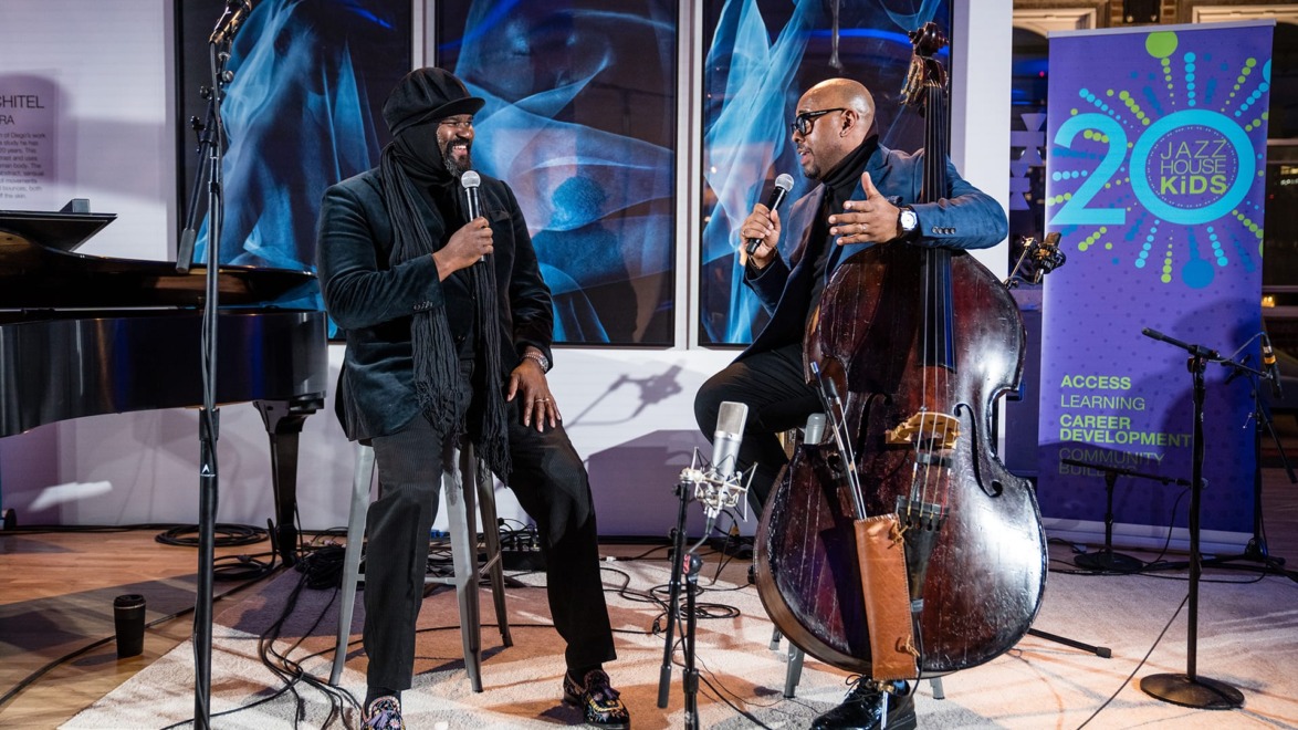 Two musicians performing with microphone and upright bass on stage, vibrant banner in background, jazz setting.