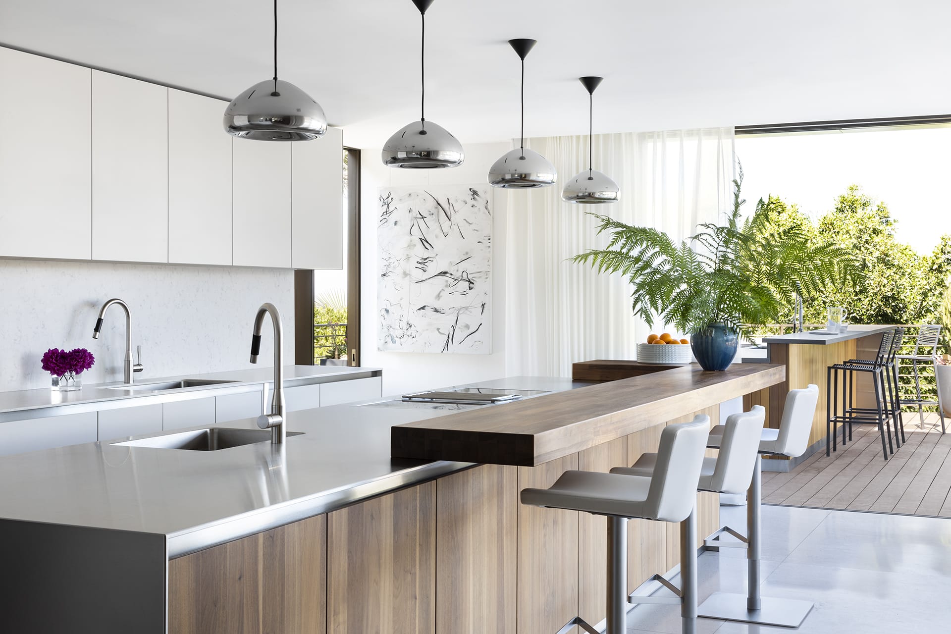 Modern kitchen with stainless steel counters, wooden island, hanging lights, bar stools, and large windows overlooking a patio.
