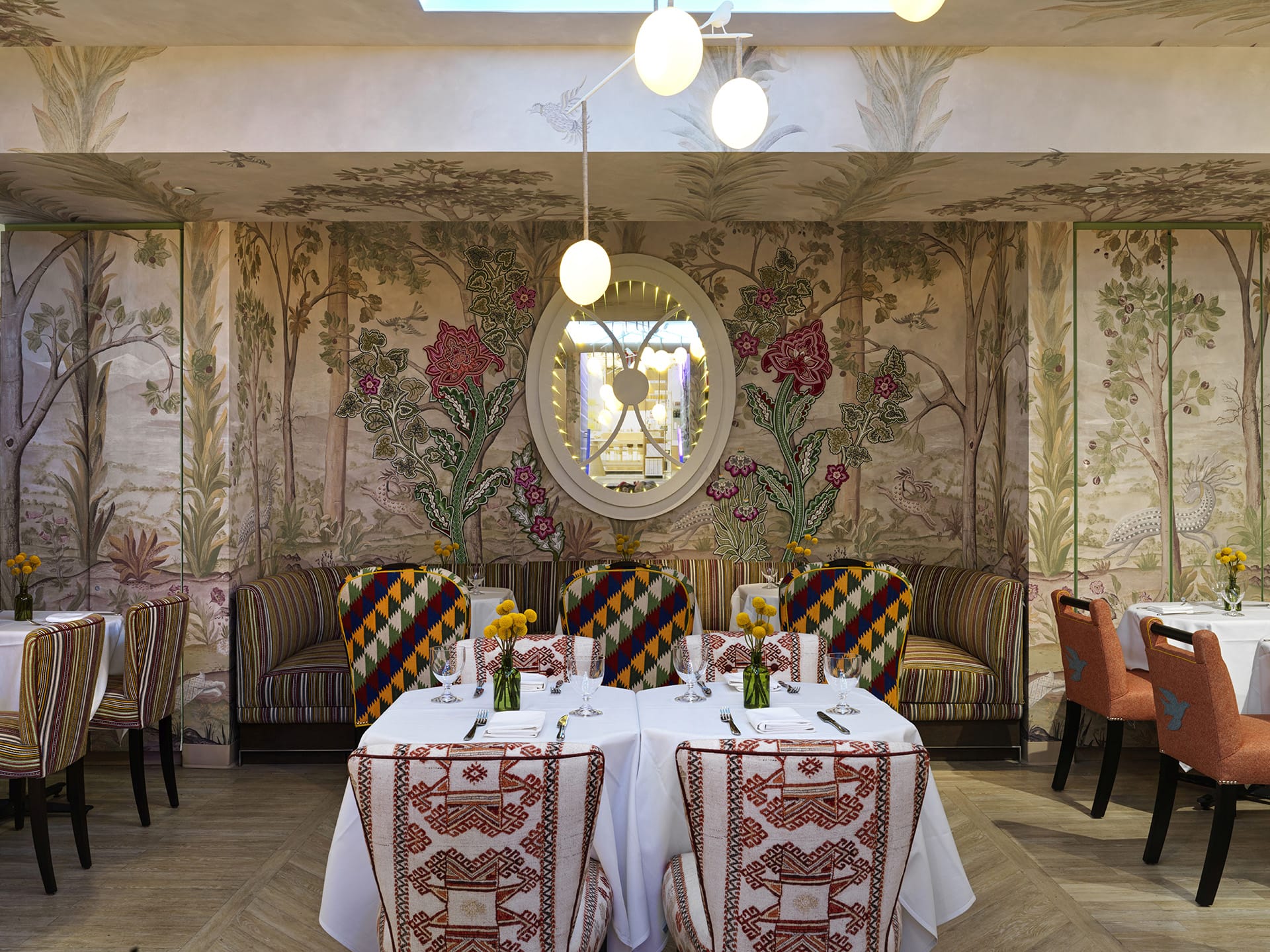 Elegant restaurant interior with floral wallpaper, round window, colorful chairs, and tables set with tablecloths and flowers.