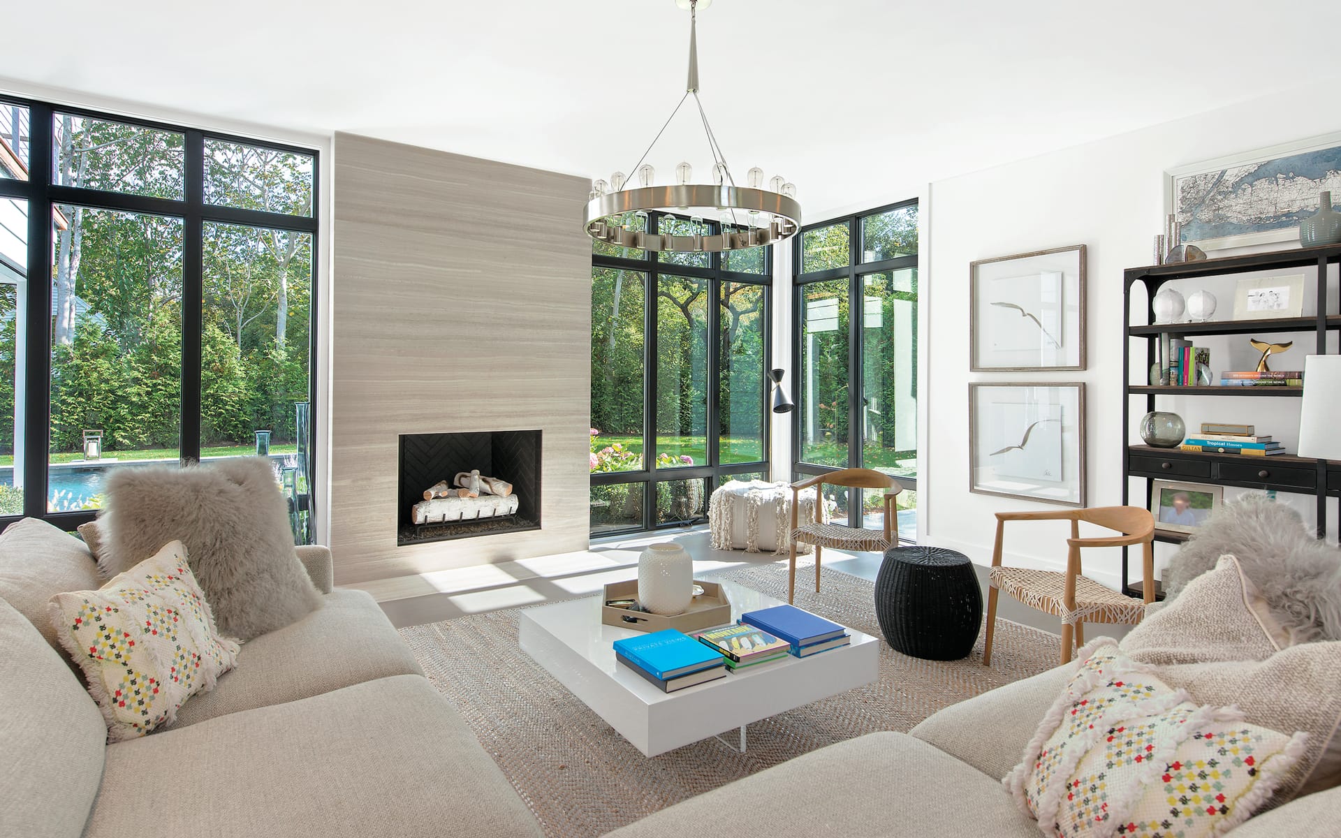 Modern living room with large windows, light furniture, a fireplace, books, and a garden view outside.
