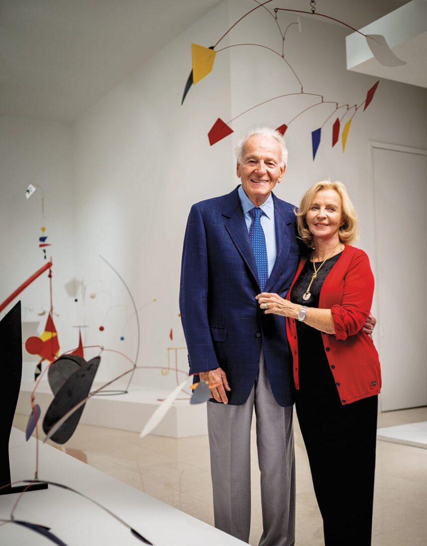 An elderly couple posing and smiling in an art gallery with colorful mobile sculptures in the background.
