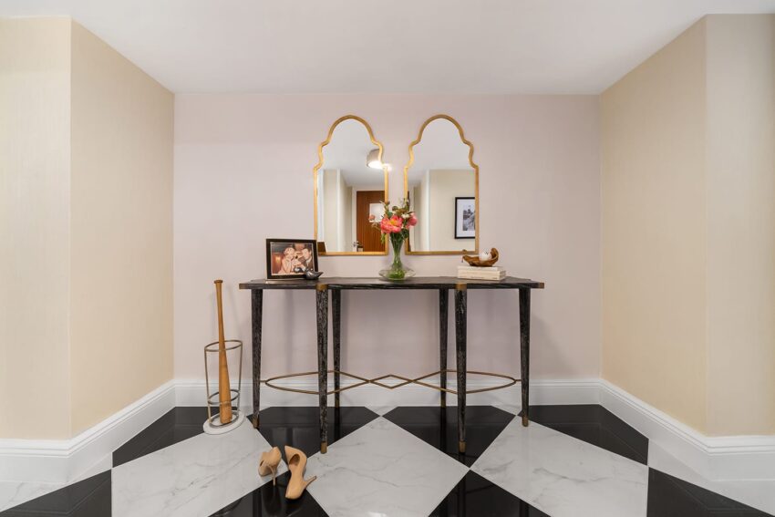 Elegant entryway with mirrored console table, fresh flowers, framed photo, and a pair of beige high heels on a checkered floor.