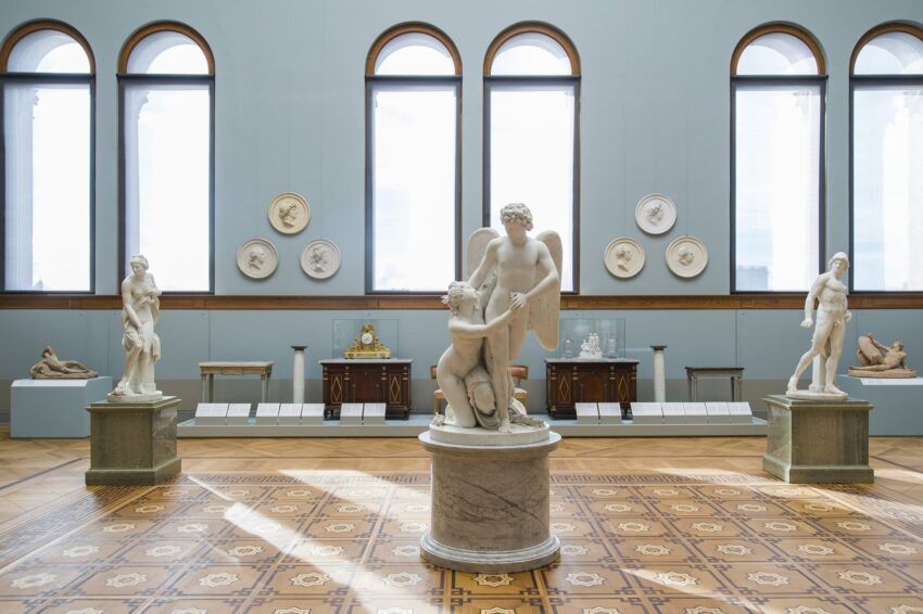 A museum room displaying various classical sculptures on pedestals with arched windows in the background.