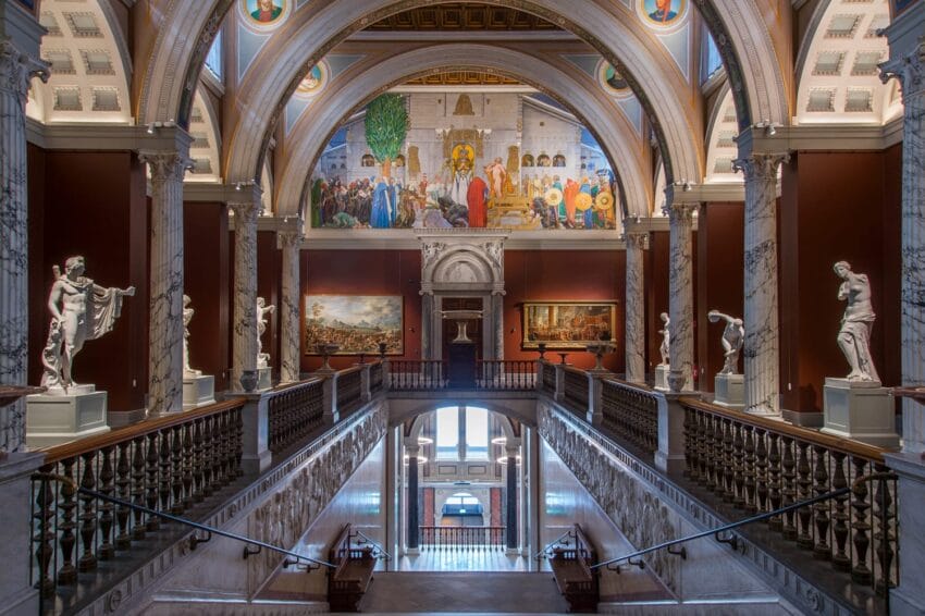 Interior of a grand museum gallery with statues, paintings, and ornate architectural details along a large staircase.