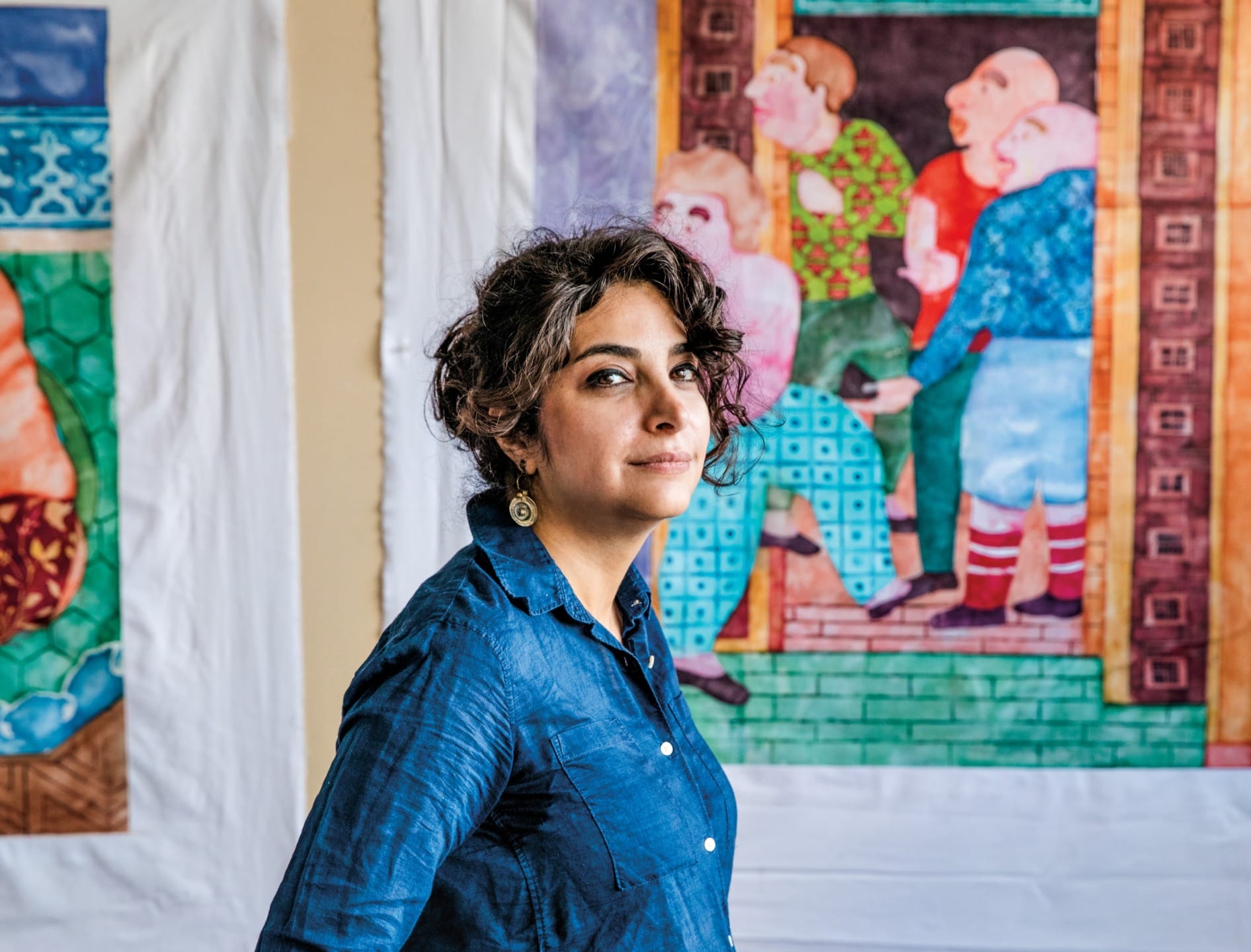 Woman in blue shirt standing in front of colorful paintings on a wall.