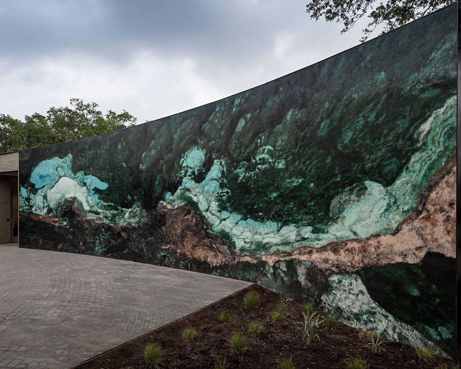 Large outdoor mural with abstract green and brown patterns on a curving wall, surrounded by trees and cloudy sky.