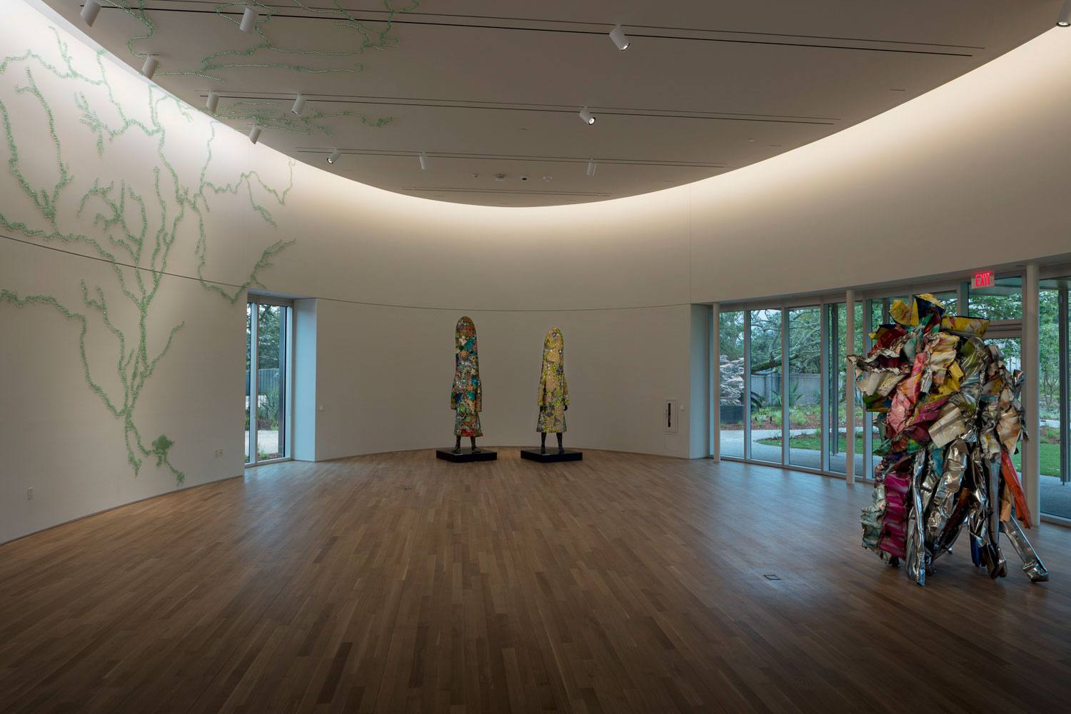 Art gallery with three colorful abstract sculptures, wooden floor, curved walls with light design, and large windows showing greenery.