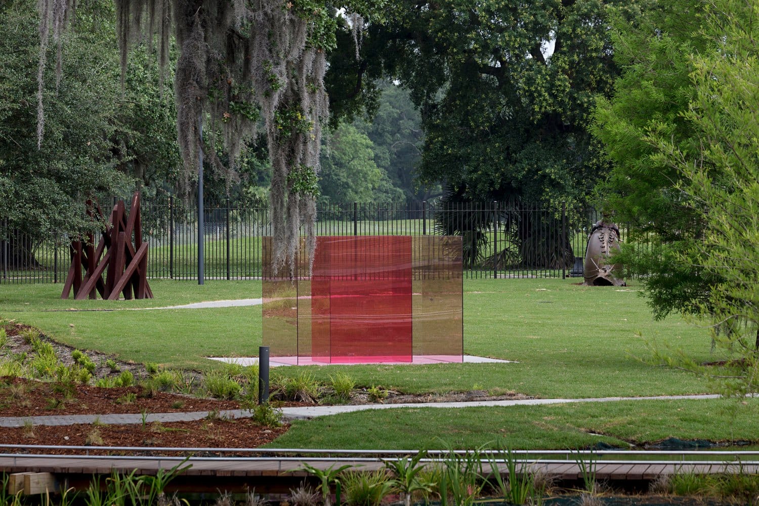 Colorful transparent sculpture in a lush garden with trees and other artworks in the background.