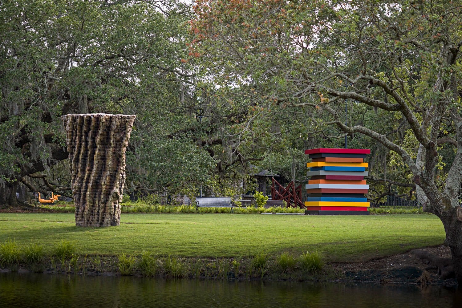 Outdoor sculpture garden with a textured column and a colorful stack, surrounded by trees and grass next to a pond.